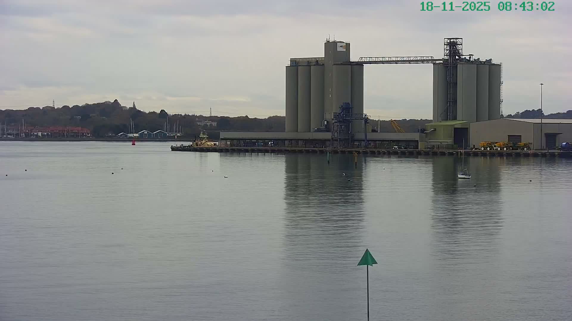 Calm grey water reflects a large industrial complex featuring silos and buildings on a pier, while a forested hillside with scattered buildings and moored boats lines the far shore under an overcast sky.