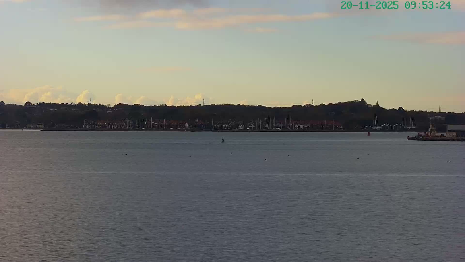 A calm body of water fills the foreground with several buoys, leading to a distant tree-lined shore featuring a coastal town with houses, a marina full of boats, and a small dock with a moored vessel, all beneath a partly cloudy morning sky.