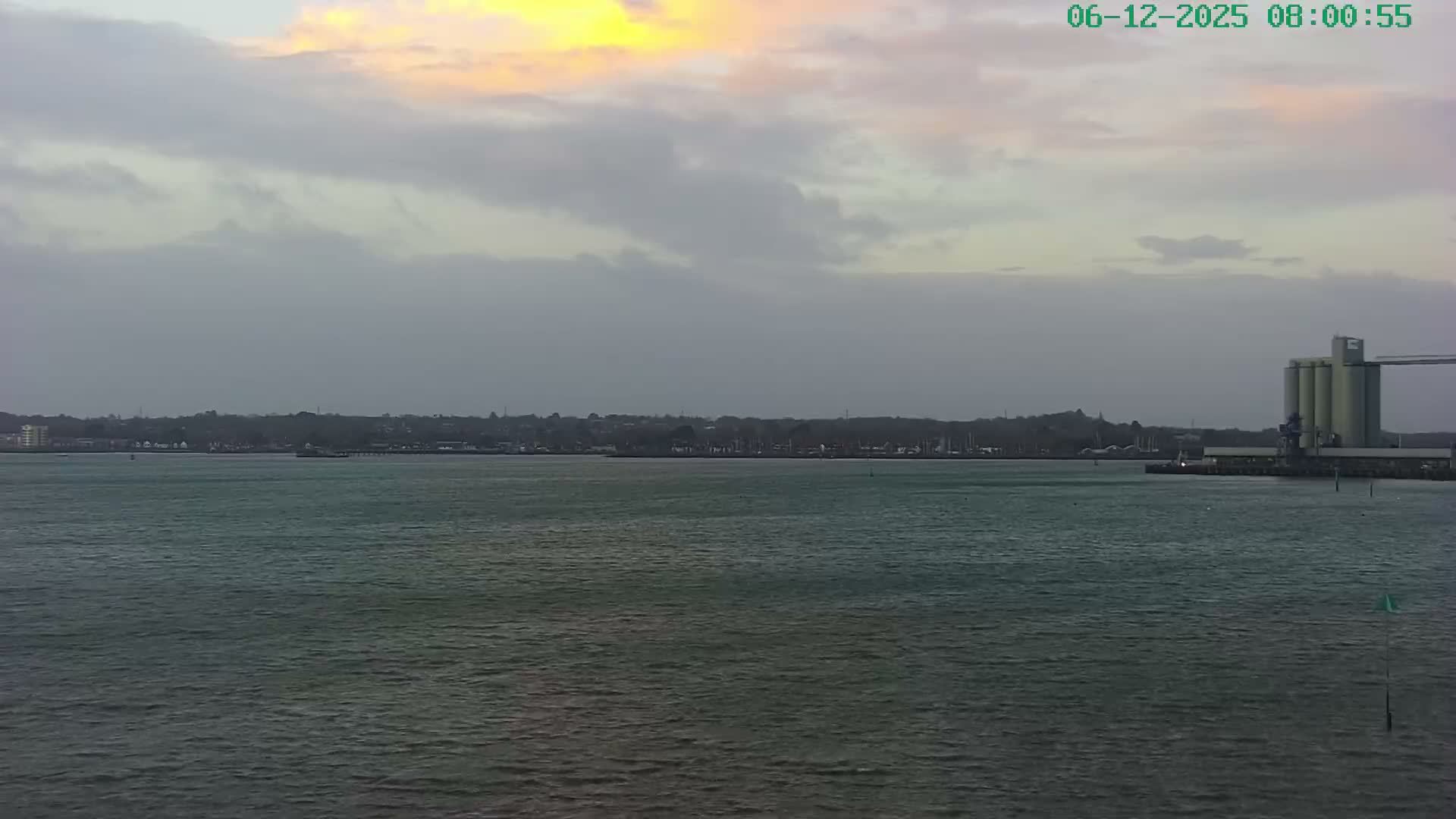 A cloudy morning with hints of sunrise illuminates a choppy body of water, bordered by a distant shoreline featuring urban buildings, a marina, and an industrial complex with large silos on the right.