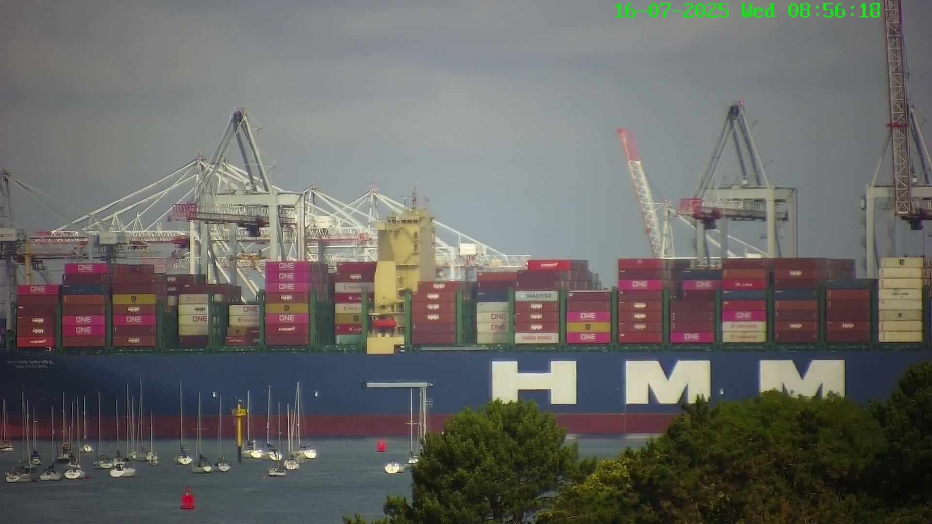 A large container ship, displaying a prominent "HMM" logo, is docked at a port under overcast skies, with numerous smaller sailboats visible in the water.