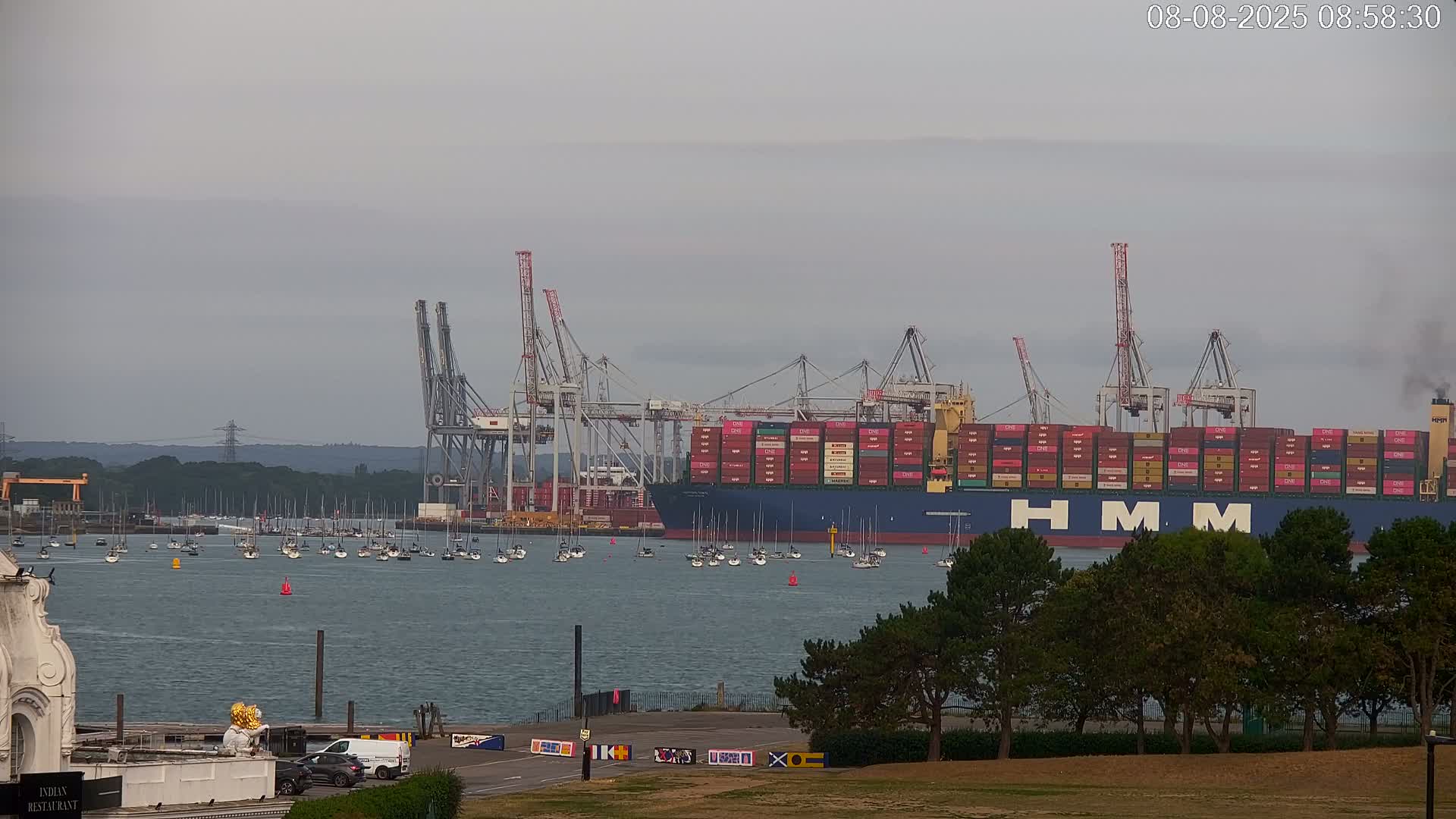 A large container ship is docked at a port under an overcast sky, with numerous smaller boats scattered in the water.