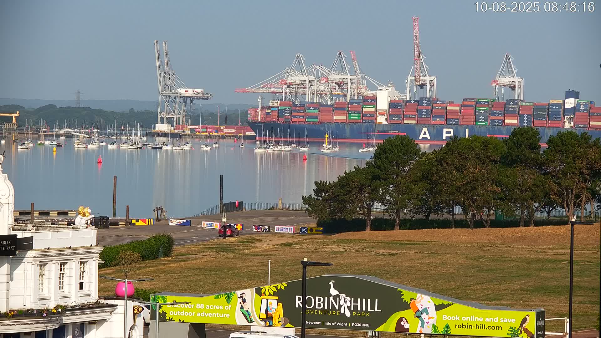 A large container ship is docked at a port, with numerous smaller boats in the water under a clear, sunny sky.
