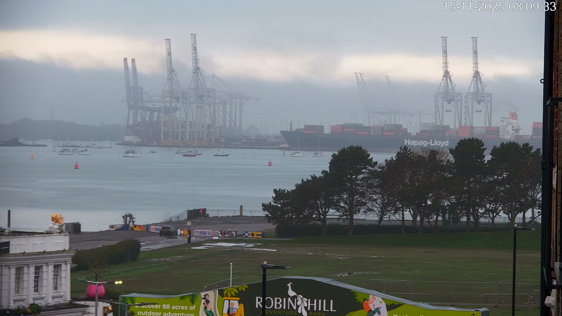 An overcast and hazy view shows a large container ship navigating a busy port with numerous cranes partially obscured by fog, alongside smaller boats on the water, with a foreground of green grass and bare trees.