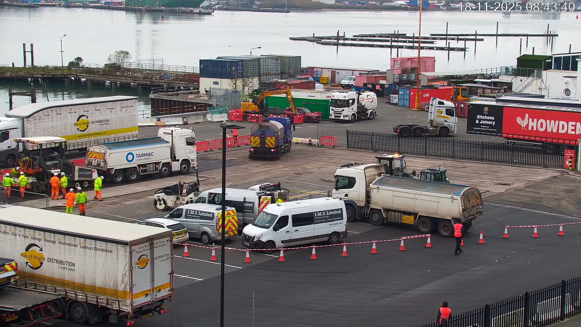 An overcast day at a bustling industrial port shows workers in high-visibility clothing tending to road paving amidst numerous trucks, vans, and shipping containers.