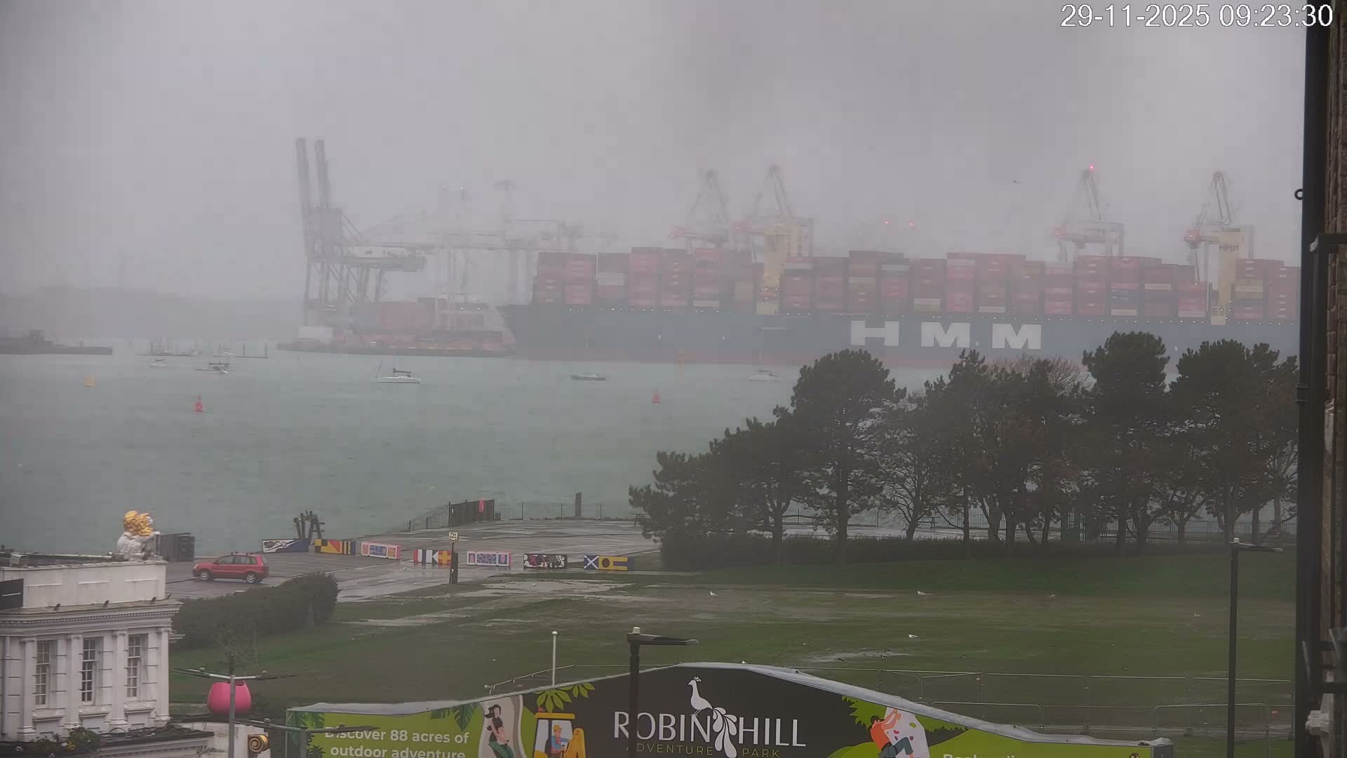 A large container ship and numerous port cranes are partially obscured by heavy rain and haze in a bustling harbor, featuring smaller boats on choppy water and a waterfront with buildings and trees in the foreground.