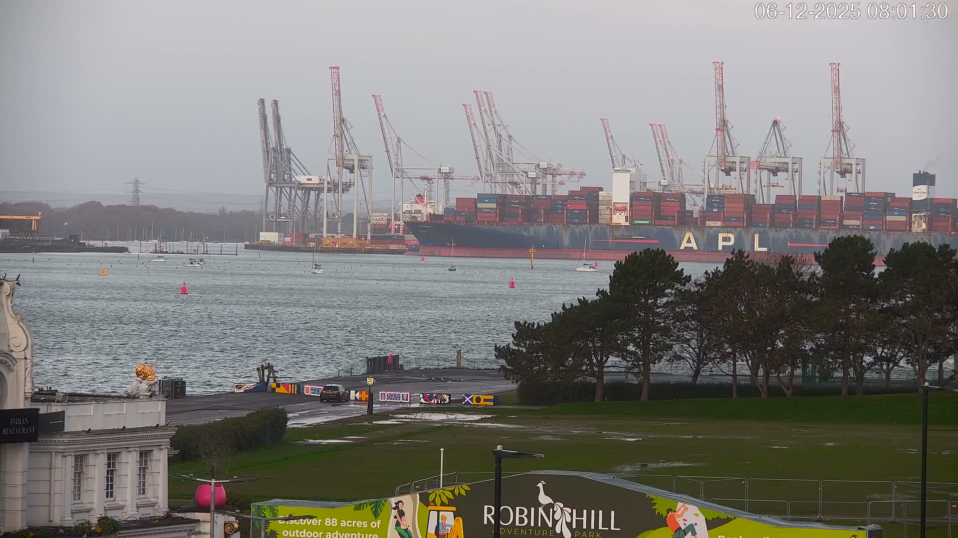 Under an overcast sky, a large container ship is docked at a busy port lined with cranes, while a choppy grey waterway dotted with small boats separates it from a wet grassy foreground featuring pine trees and a section of a white building.