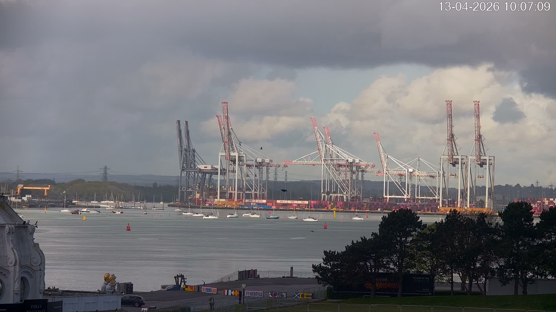 Under an overcast sky, a large container ship is docked at a busy port lined with cranes, while a choppy grey waterway dotted with small boats separates it from a wet grassy foreground featuring pine trees and a section of a white building.