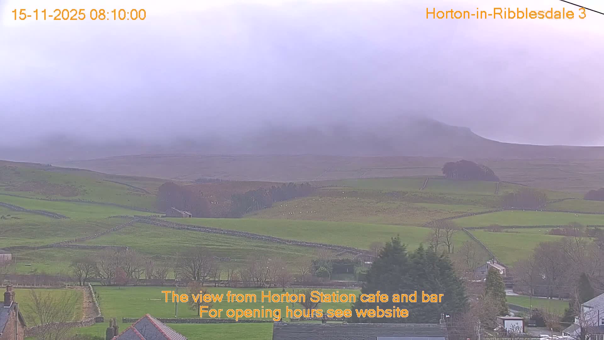 A misty morning view reveals a rural landscape of green fields separated by stone walls, with bare trees and distant hills fading into heavy fog under an overcast sky.