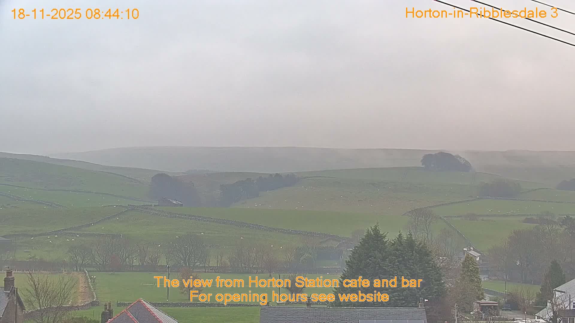 The image displays a misty, overcast rural landscape featuring rolling green hills sectioned by stone walls, scattered bare trees, and rooftops of a few buildings in the foreground.