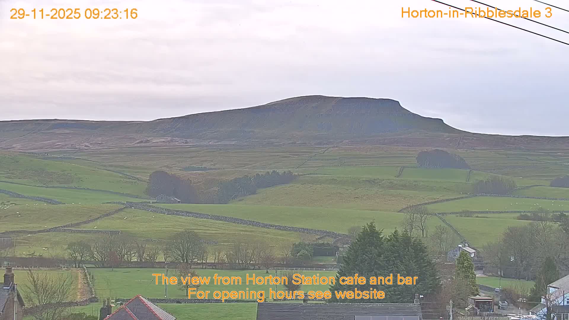 Under an overcast grey sky, a vast rural landscape displays rolling green fields divided by numerous stone walls, leading up to a large, barren hill in the distance, with parts of a village visible in the foreground.