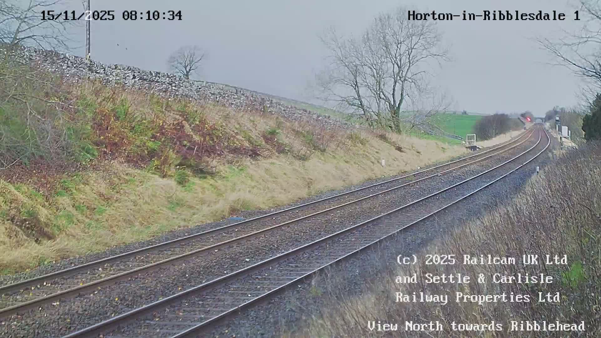 A double-track railway line extends through a rural, hilly landscape with dry stone walls and bare trees under an overcast sky, curving into the distance towards a distant bridge and a red signal.
