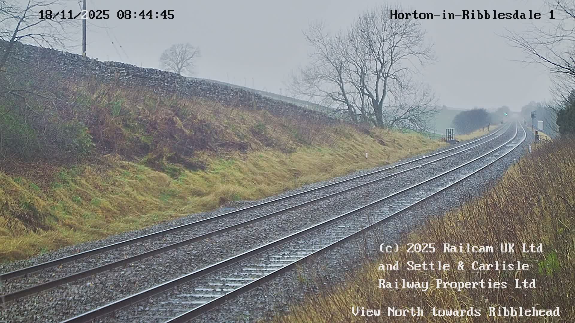 The image shows a double-track railway curving into the distance between grassy, autumnal hillsides and bare trees, under a very overcast and misty sky.