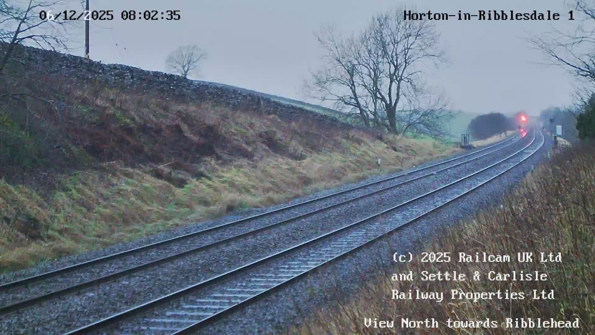 Under a dull, overcast sky, two railway tracks stretch into a misty distance between grassy hills and bare trees, with faint red lights visible on the horizon.