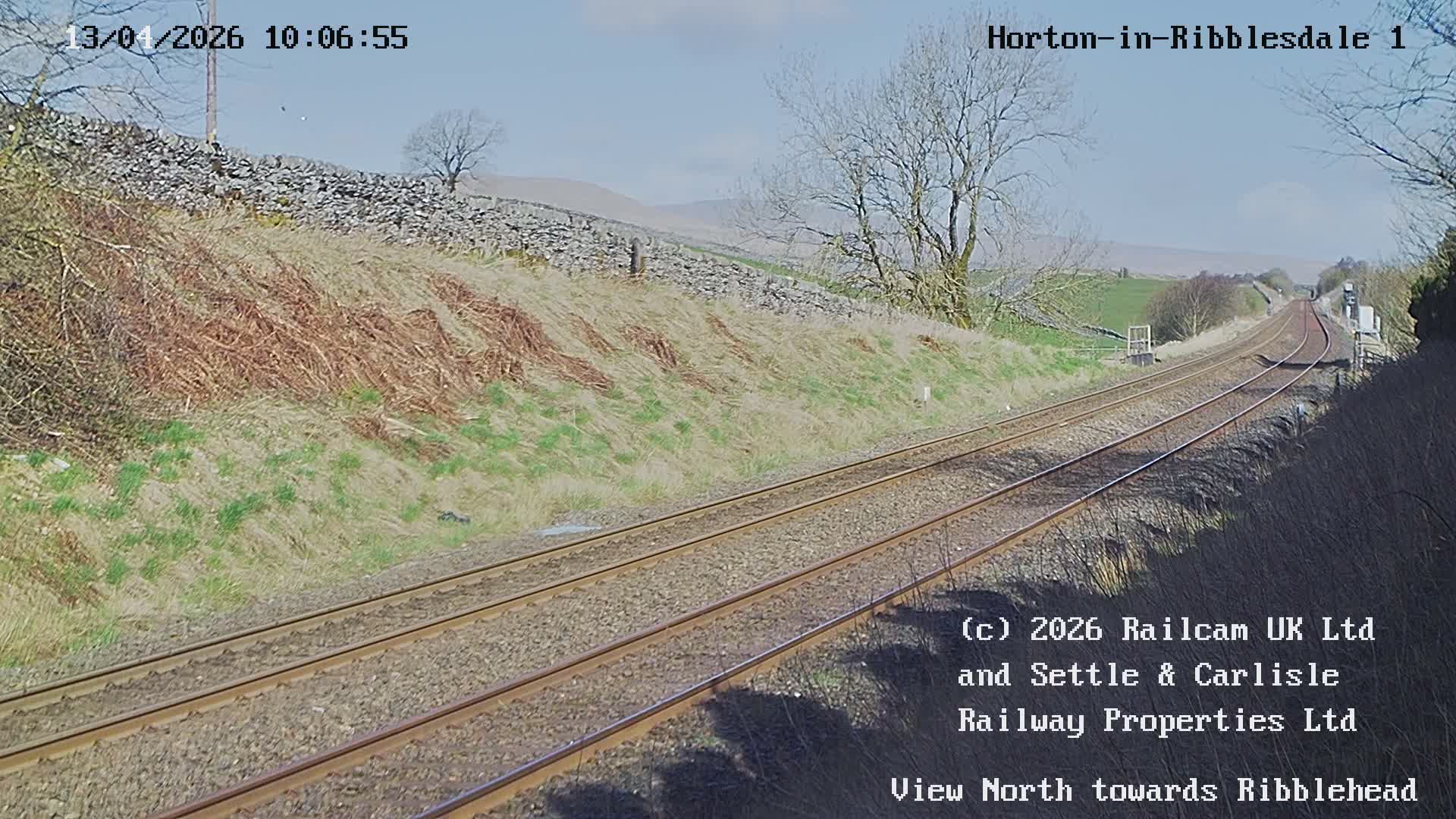 Under an overcast sky, a double railway track curves through a rural landscape with brown grassy slopes and bare trees, leading to distant red signal lights.
