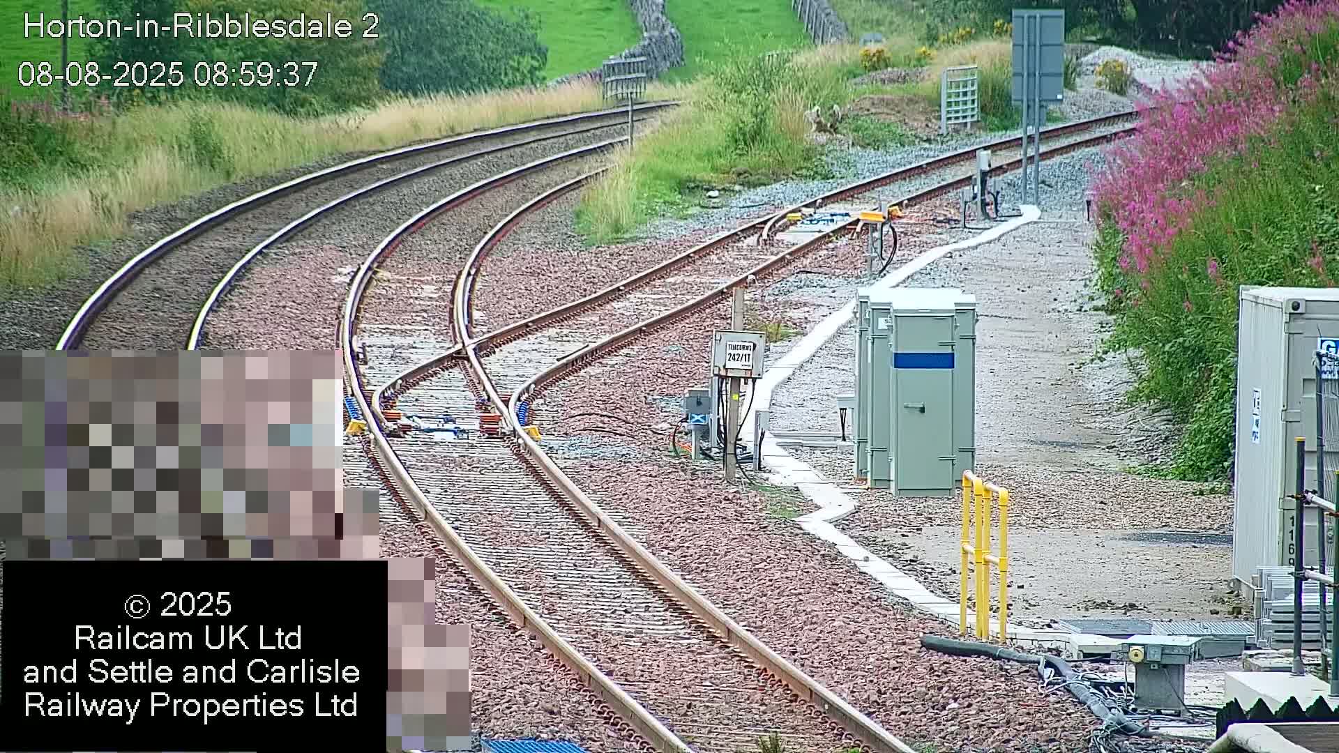 A curving railway track with several parallel lines runs alongside a gravel path bordered by vegetation and some utility boxes, all under an overcast sky.