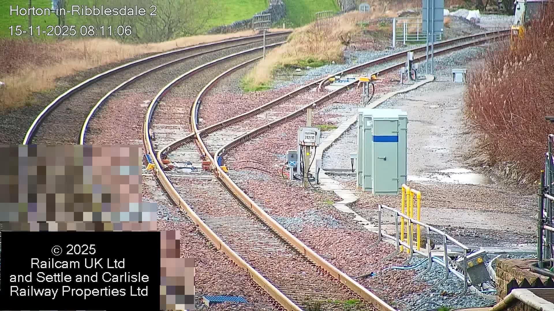 Multiple railway tracks curve and converge across a gravel bed, flanked by grassy slopes and dry, brownish bushes with utility boxes and puddles on a nearby path, under overcast and damp conditions.