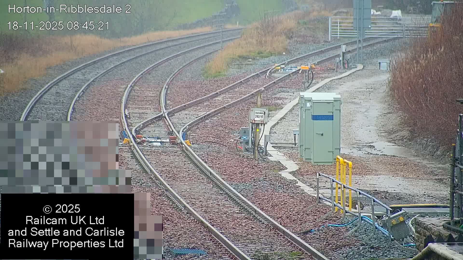 A complex network of railway tracks with switches and signal equipment lies on a gravel bed, bordered by sparse, brown vegetation and muddy patches, all under overcast and damp conditions.