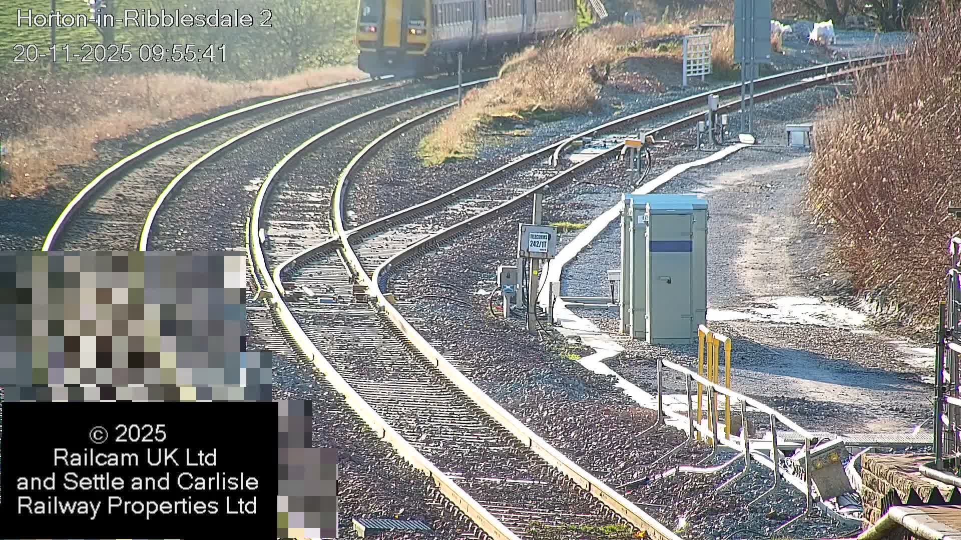 On a clear, sunny day, a yellow and grey train moves through a railway junction featuring multiple tracks, switches, and trackside utility cabinets.