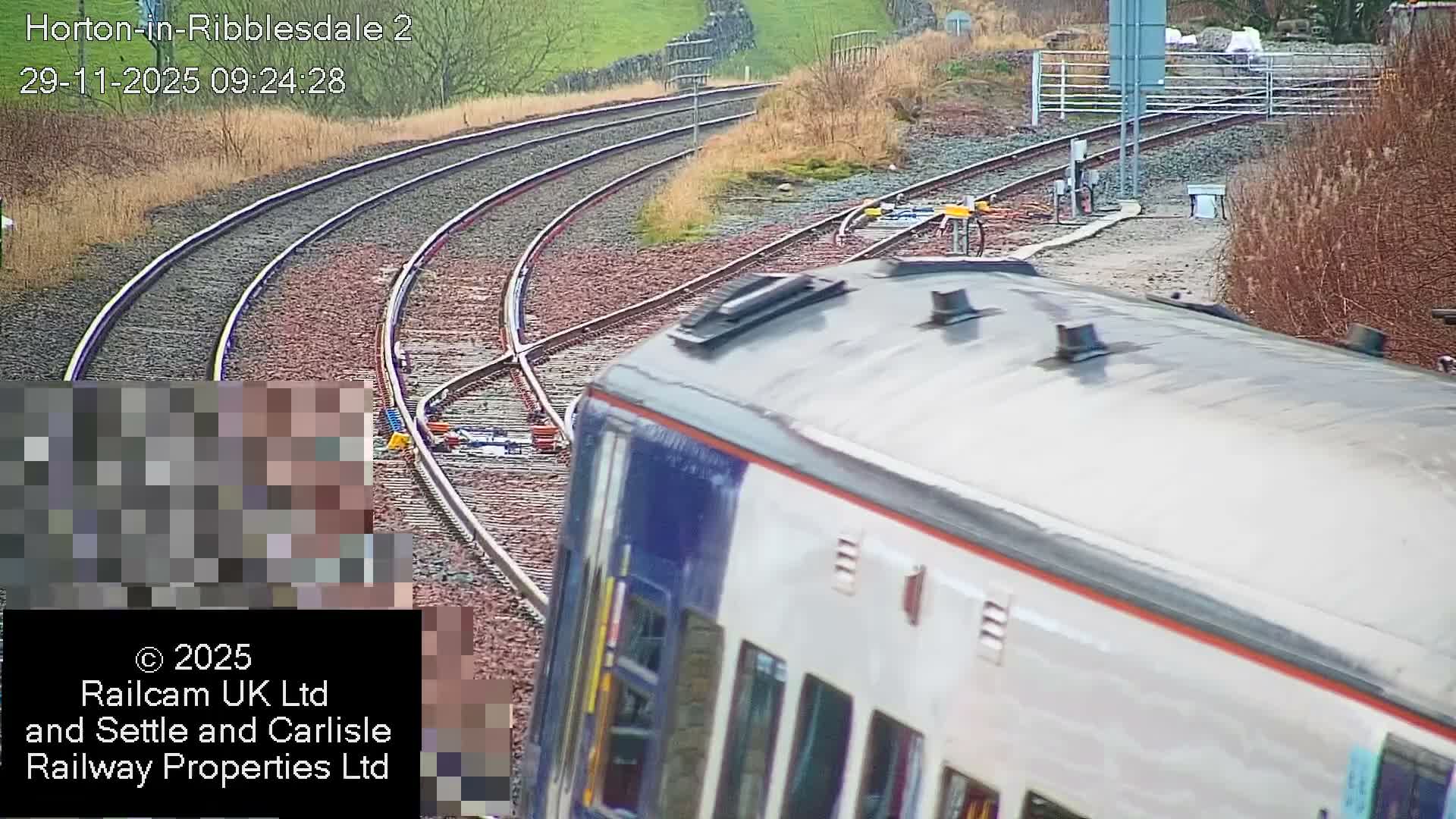 A blue and white train is seen from an elevated perspective passing along multiple railway tracks that curve through a green and brown rural landscape under overcast skies.