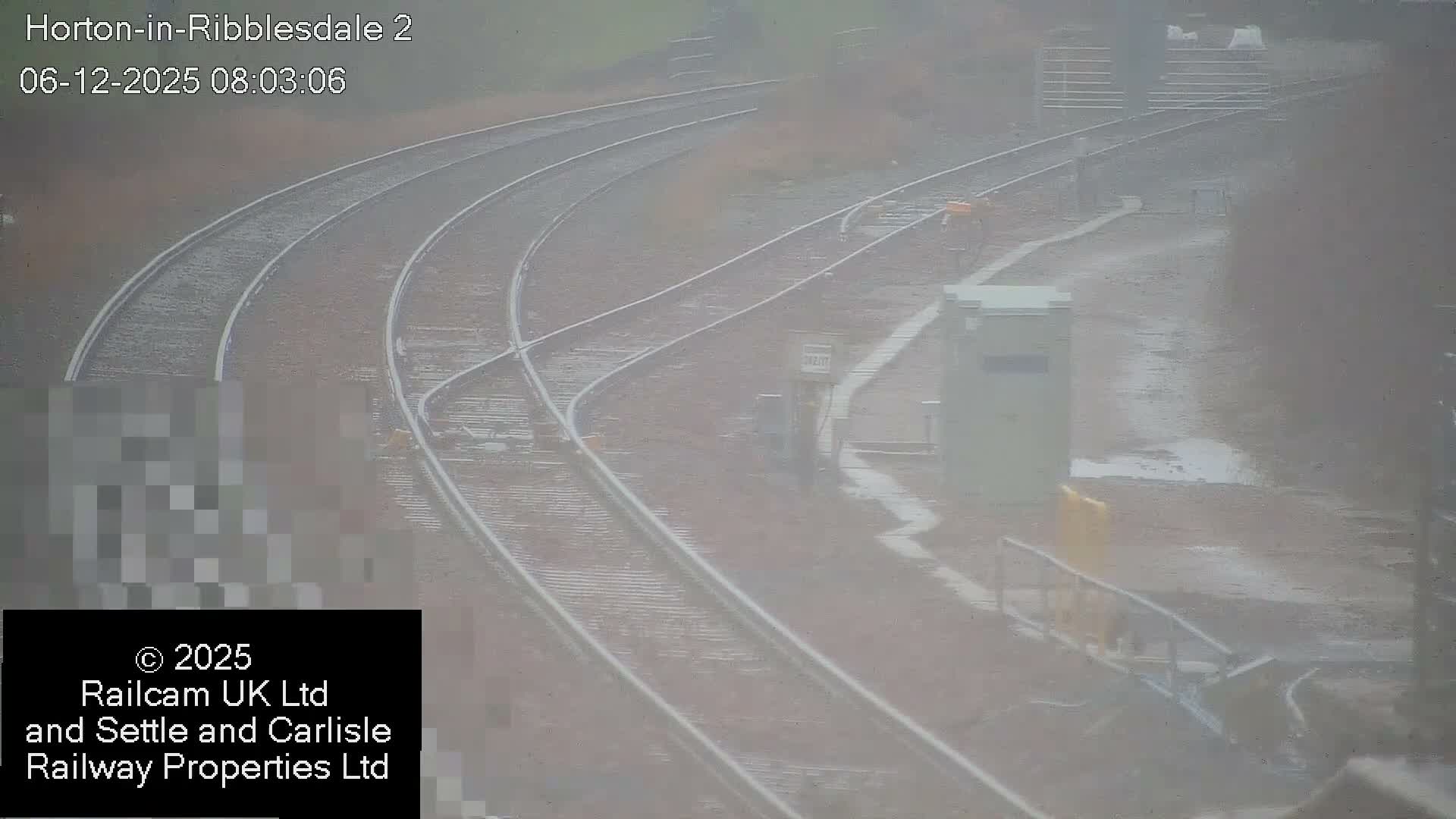 Several railway tracks curve and converge on a misty, overcast day with wet ground and puddles alongside various railway infrastructure.