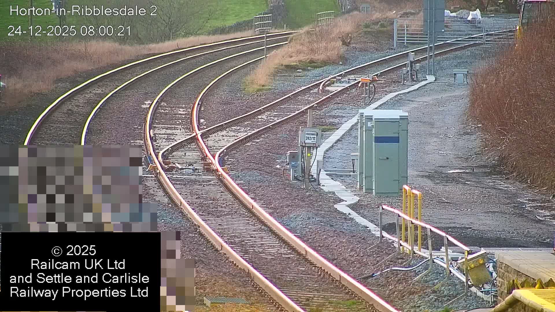 Multiple railway tracks converge and diverge at a rural junction with signal boxes and equipment, set against grassy hills and bare bushes under clear, dry conditions.