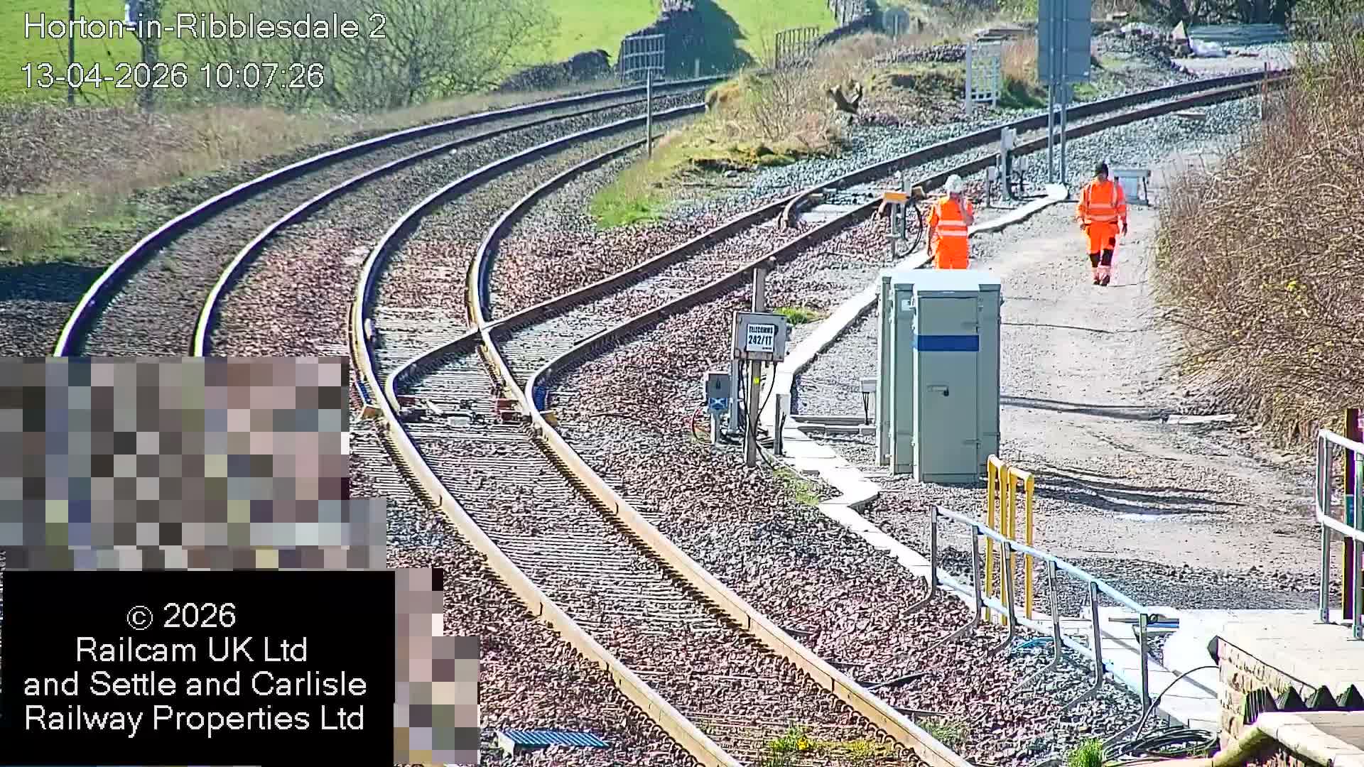 Multiple railway tracks converge and diverge at a rural junction with signal boxes and equipment, set against grassy hills and bare bushes under clear, dry conditions.