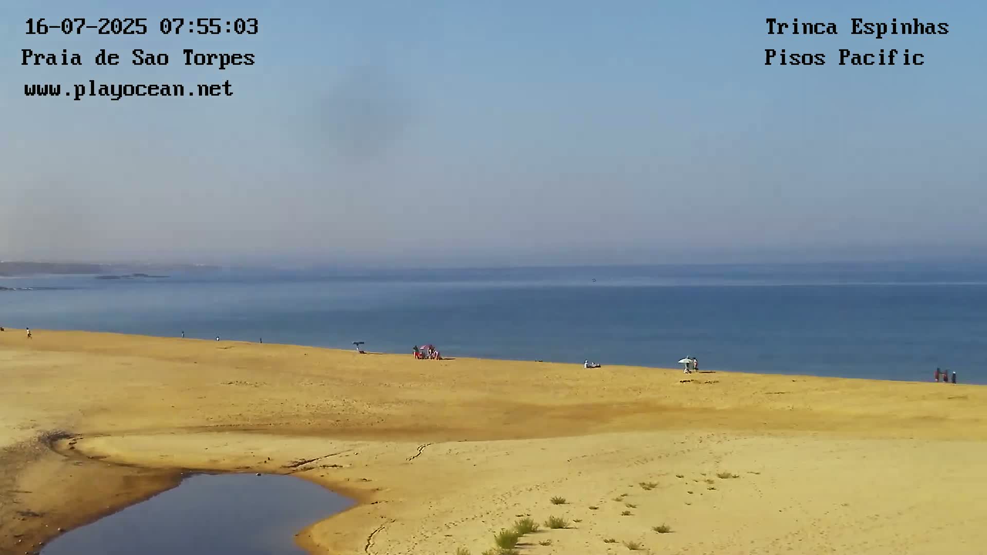 A sandy beach with a few people and umbrellas stretches along a calm blue ocean under a clear sky.