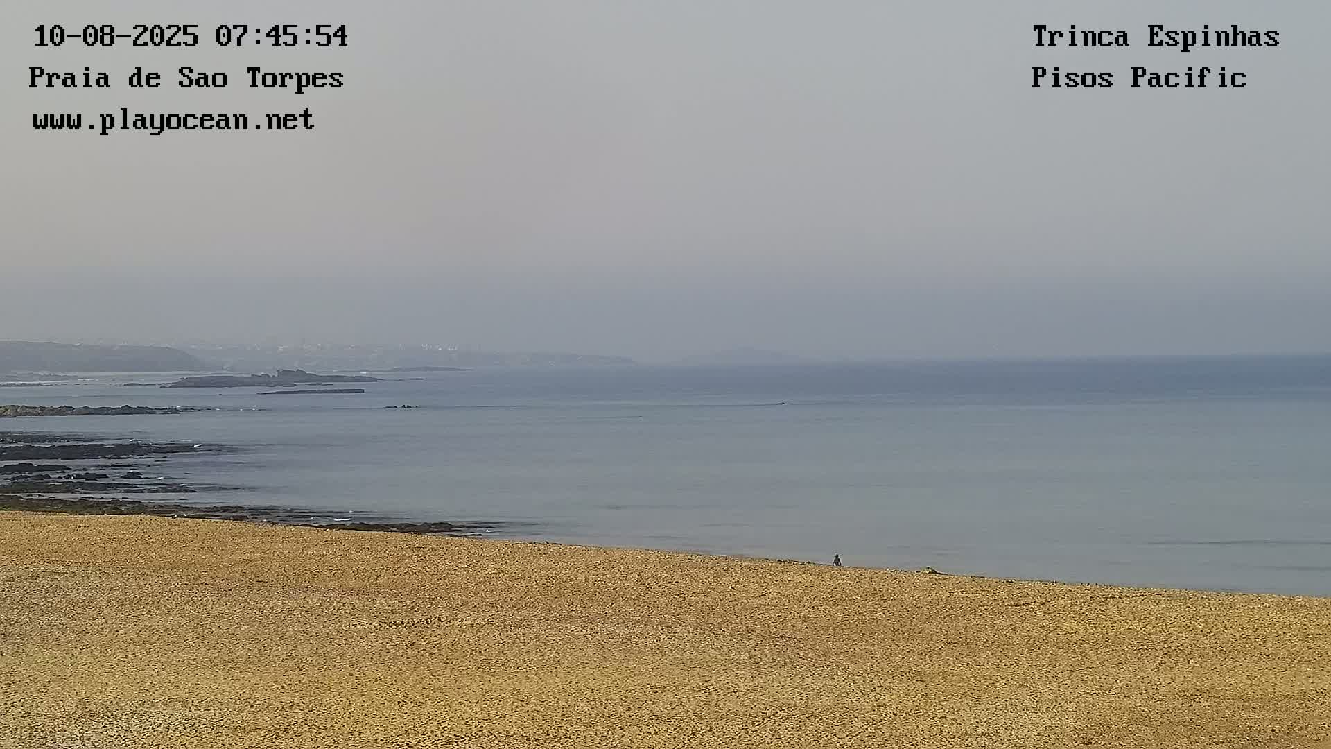 A hazy day shows a long sandy beach sloping down to a calm ocean with dark rocks and distant land visible.