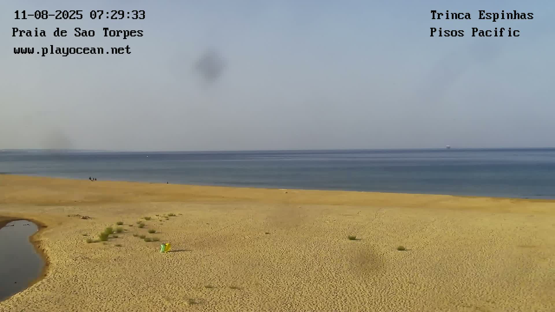 A mostly clear sky overlooks a long, sandy beach and calm ocean, with a few people visible on the sand.
