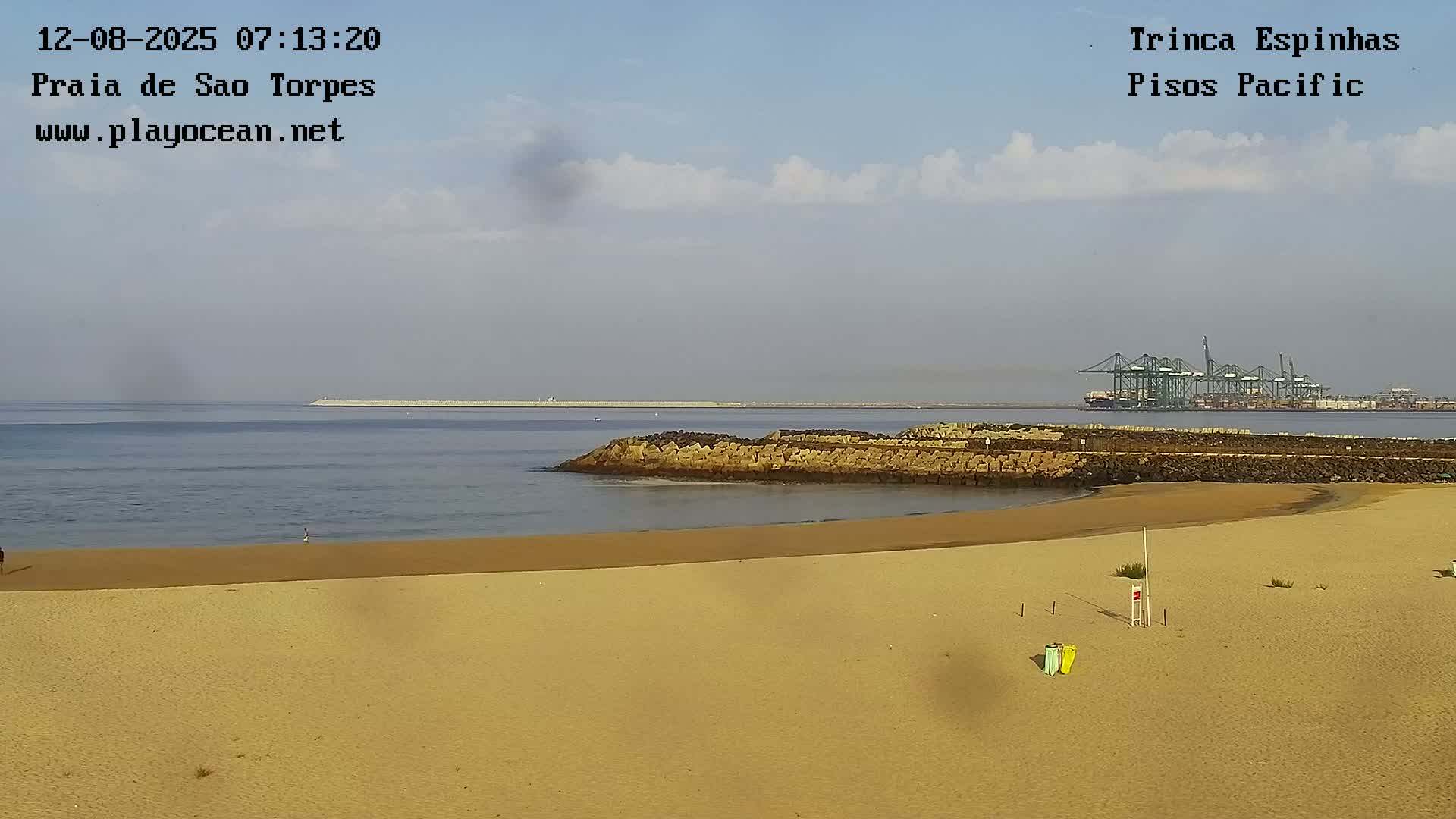 A sandy beach with calm water, a rocky breakwater, and a distant port with large cranes under a partly cloudy sky.