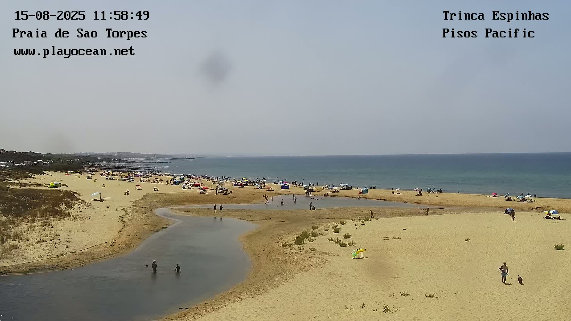 A sandy beach with calm water, a rocky breakwater, and a distant port with large cranes under a partly cloudy sky.