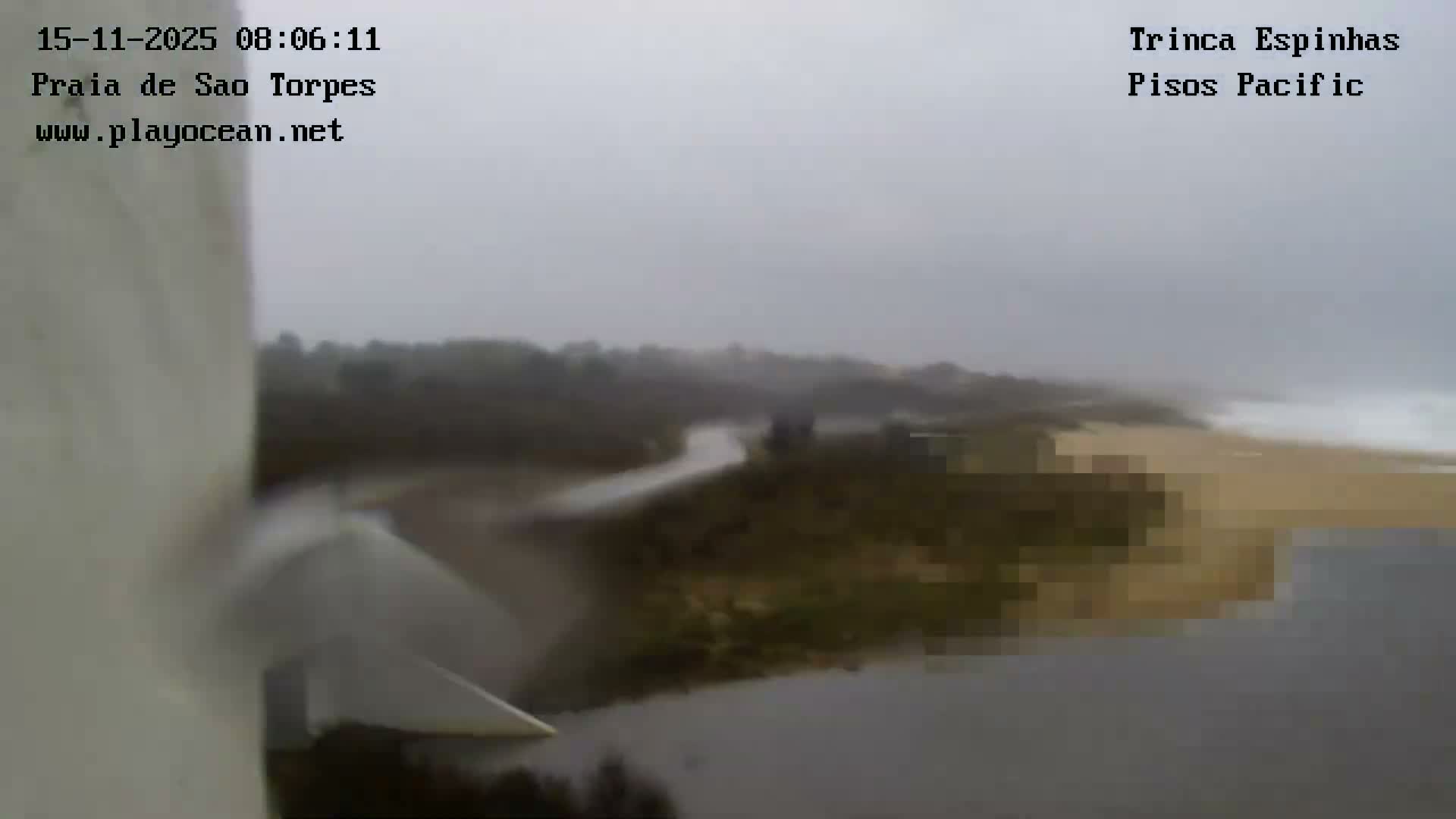 A heavy overcast sky covers a coastal landscape featuring a river opening onto a sandy beach with breaking waves, while a large white structure partially frames the left foreground.