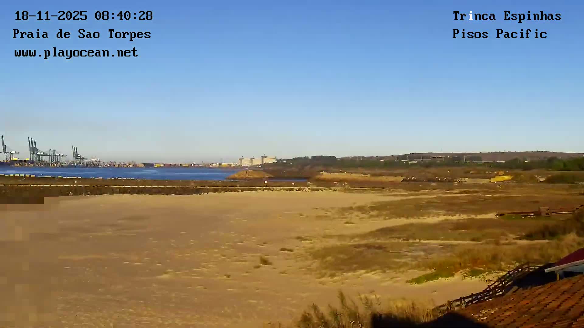A heavy overcast sky covers a coastal landscape featuring a river opening onto a sandy beach with breaking waves, while a large white structure partially frames the left foreground.