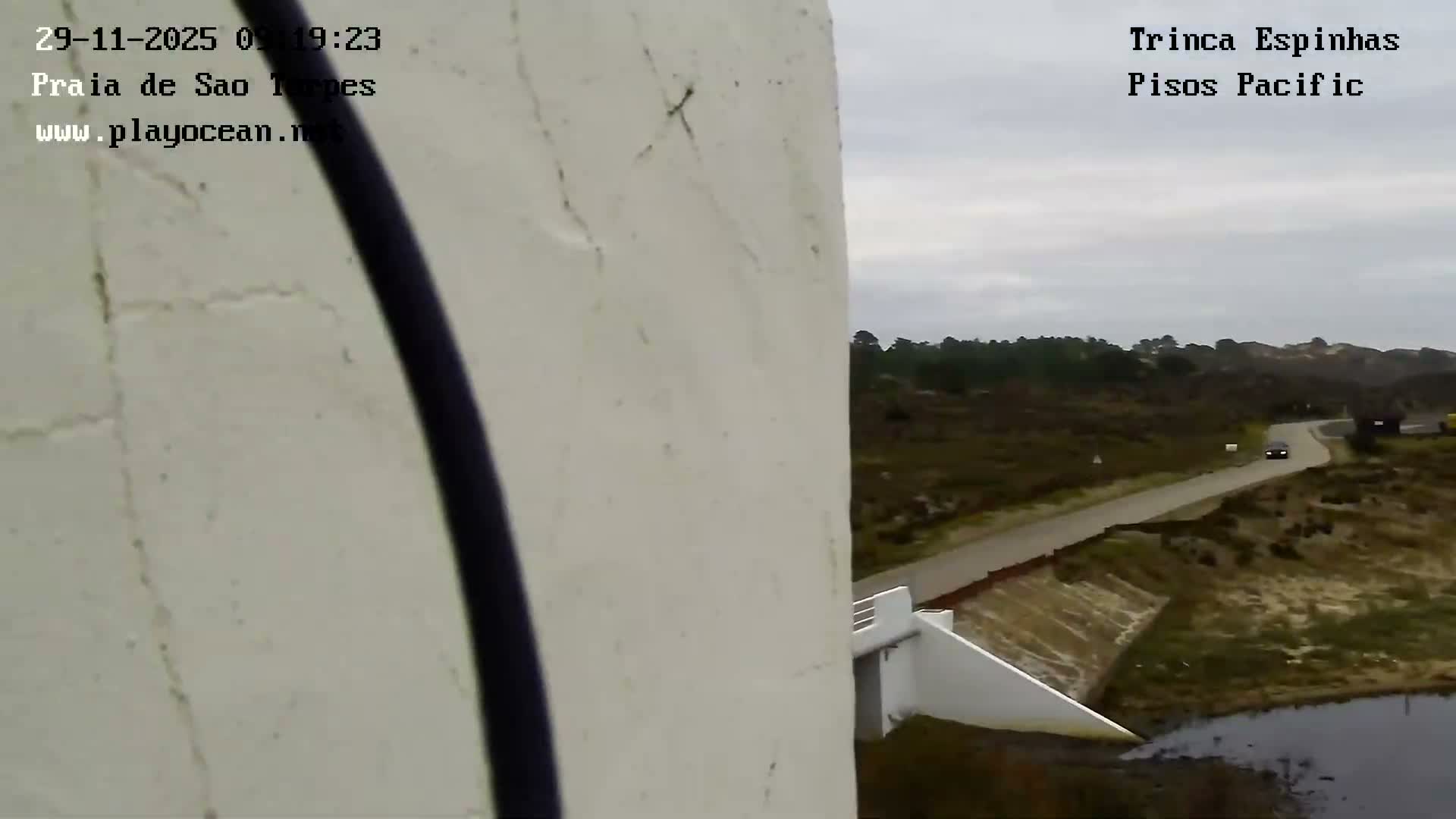 Under an overcast sky, a dark car drives along a winding road through a sparsely vegetated, hilly landscape with a small body of water, partially framed on the left by a light-colored wall and a vertical dark cable.