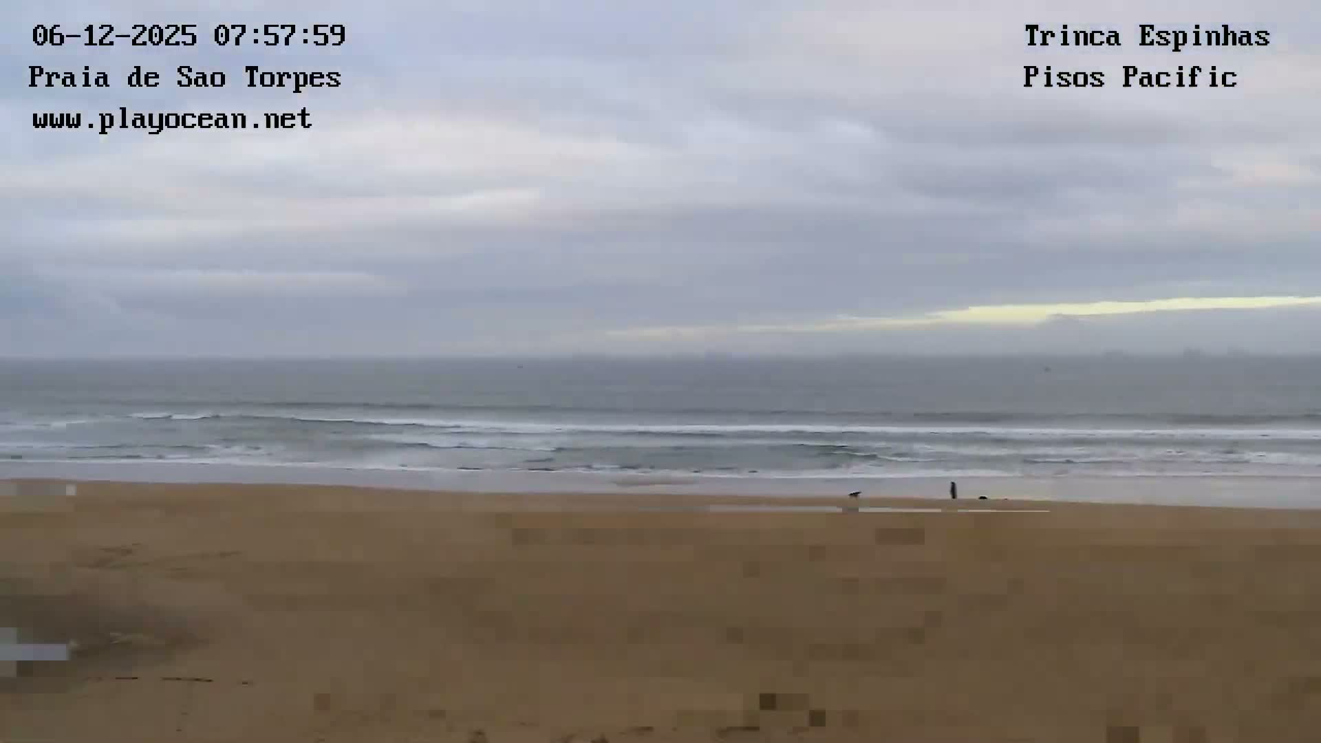 A wide sandy beach stretches towards an ocean with breaking waves under a cloudy, overcast sky, with a few distant figures visible near the water's edge.