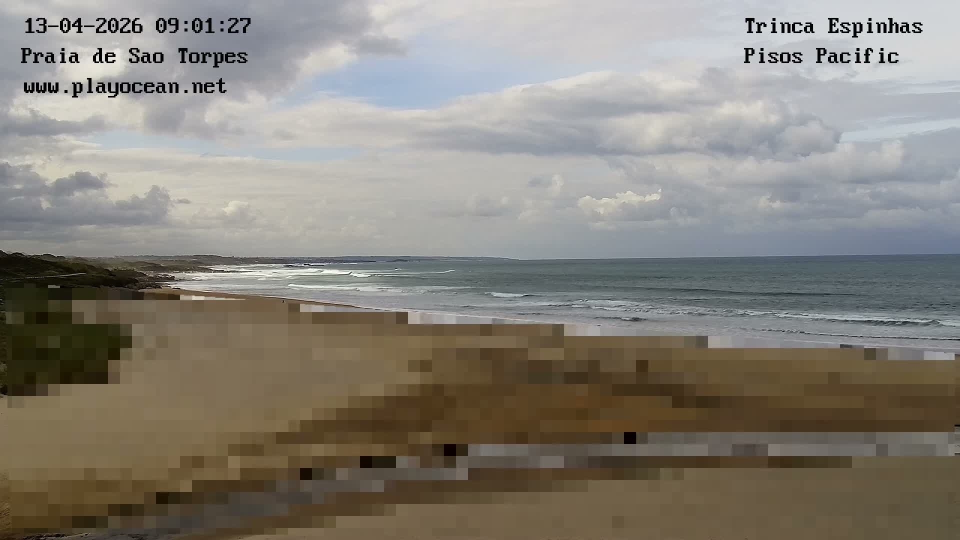 A wide sandy beach stretches towards an ocean with breaking waves under a cloudy, overcast sky, with a few distant figures visible near the water's edge.