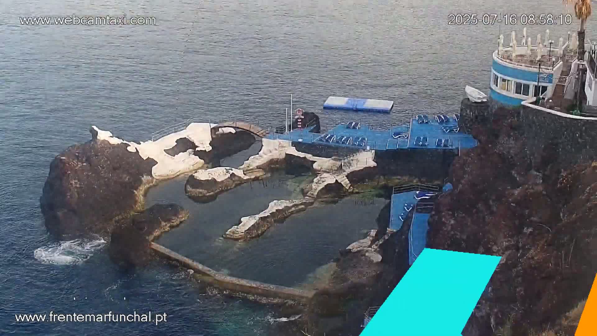 An oceanside pool complex built into dark volcanic rock, with a small building nearby, on a partly sunny day.
