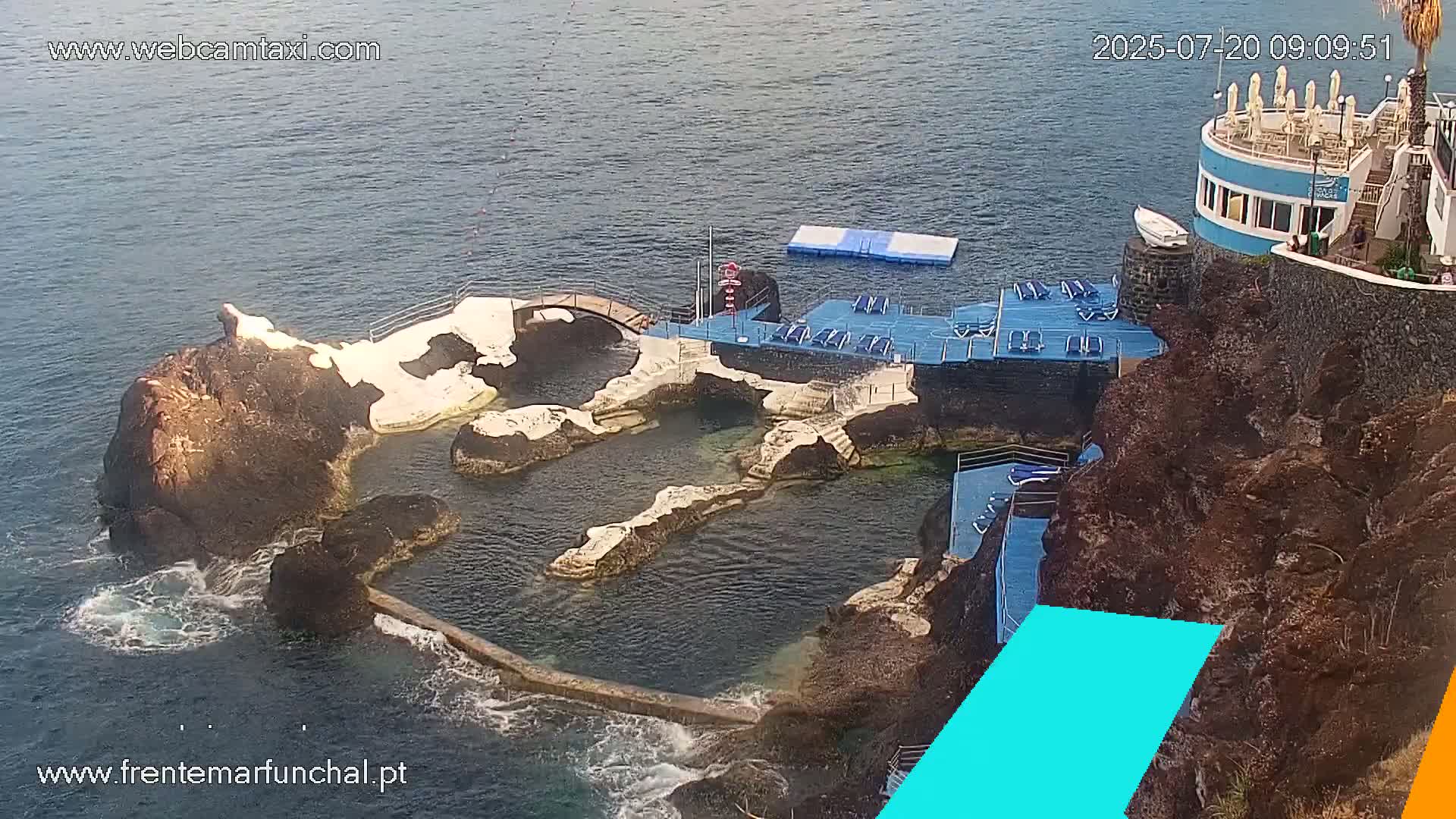 An outdoor aerial view shows a rocky coastal area with multiple pools, a diving platform, lounge chairs, and a building on a cliff overlooking calm ocean water.