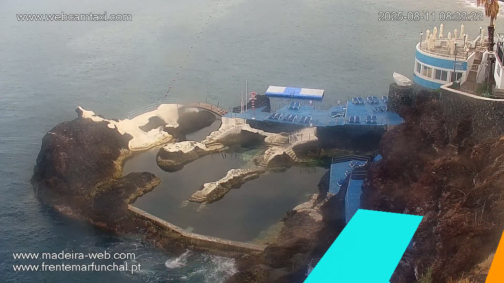 An aerial view shows a rocky coastal area with a man-made pool complex and a circular building, all under hazy or overcast conditions.