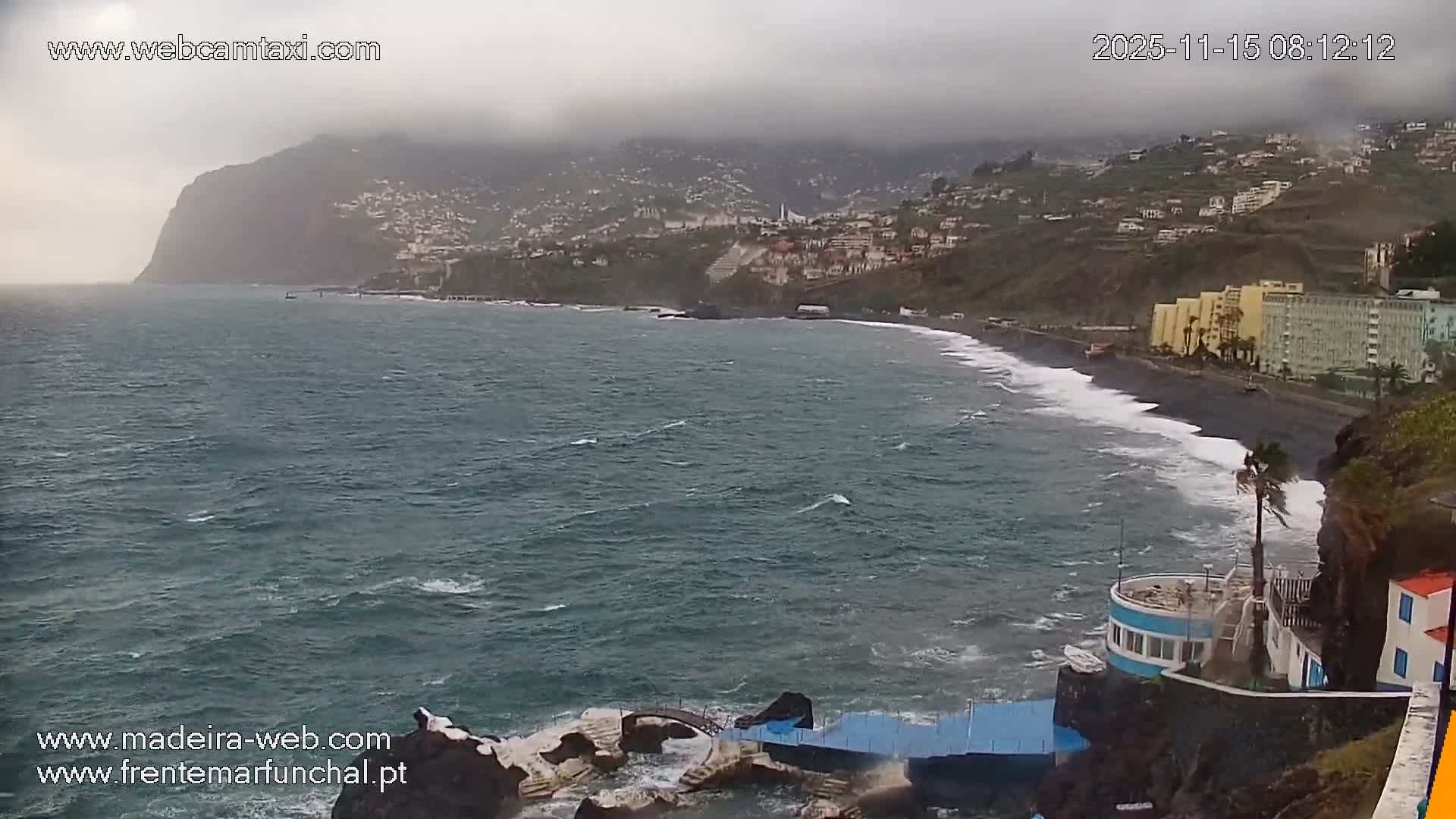 Under an overcast and cloudy sky, rough turquoise waves crash against a black sand beach and coastal structures, including lidos, with numerous buildings climbing the steep hillsides.