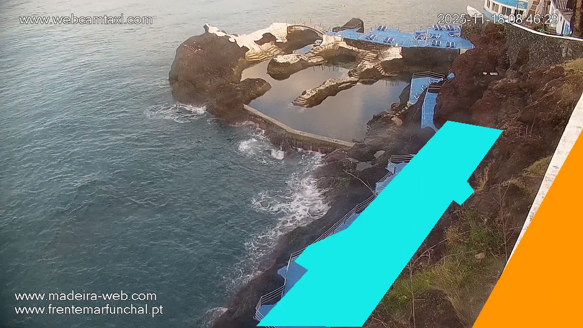 An aerial view captures a rocky coastline featuring a series of natural sea pools and a sunbathing platform with blue lounge chairs, adjacent to the choppy ocean under bright, clear weather.