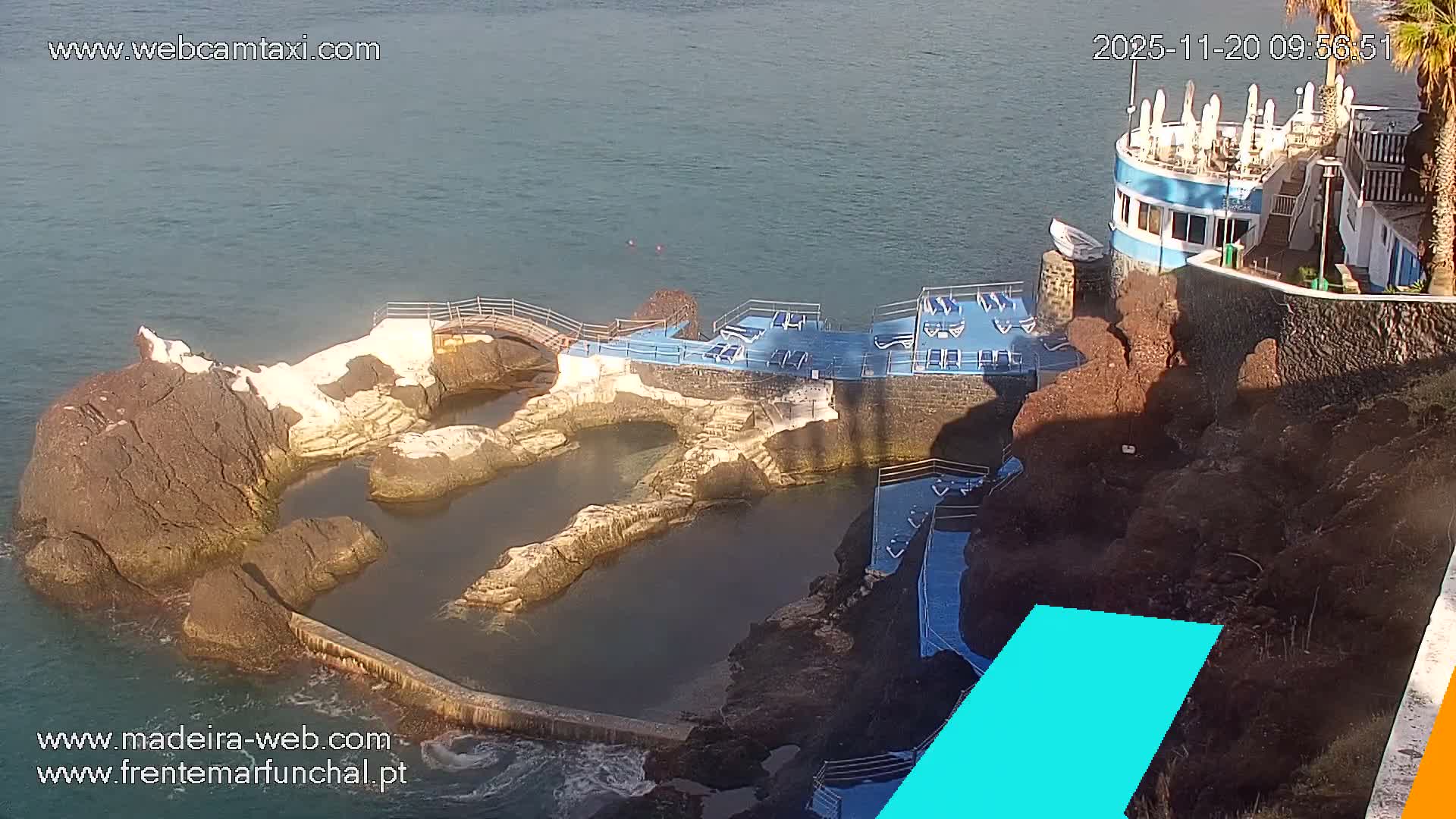 On a bright, sunny day, a coastal view captures natural ocean-fed rock pools, a blue terraced sun deck with lounge chairs, and a multi-level building with umbrellas situated along a rocky shore beside the calm sea.