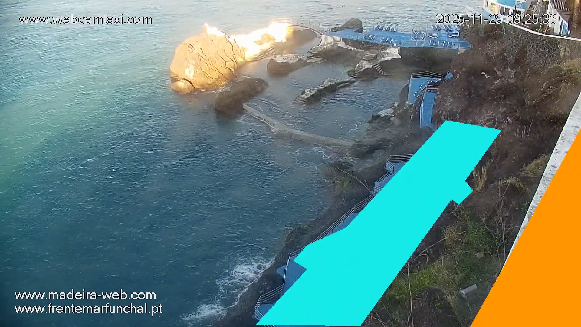 The image captures a sunny coastal landscape with calm blue-green ocean water, natural rock formations, and a multi-level leisure area featuring blue-railed stairs and platforms with sun loungers built into a steep, vegetated rocky cliffside.