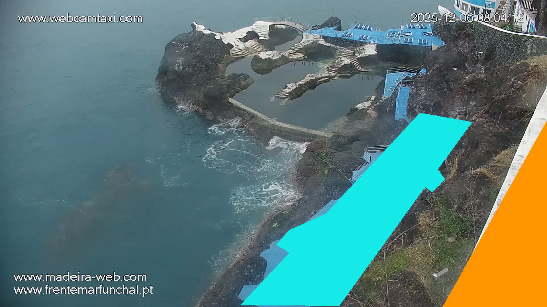 A rugged coastline features a series of natural rock pools integrated with concrete walkways, steps, and blue lounge platforms, all overlooking an ocean with moderate waves under what appears to be an overcast sky.