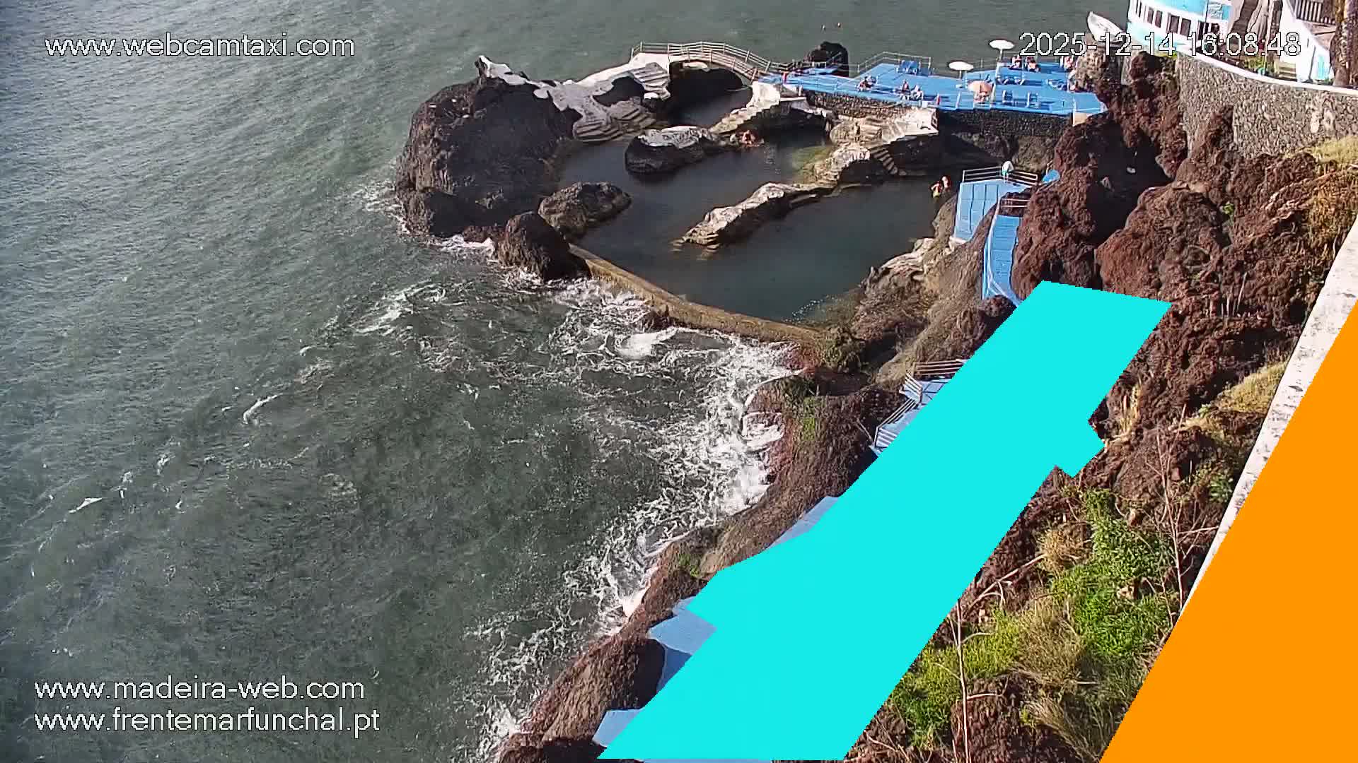 A rugged coastline features a series of natural rock pools integrated with concrete walkways, steps, and blue lounge platforms, all overlooking an ocean with moderate waves under what appears to be an overcast sky.
