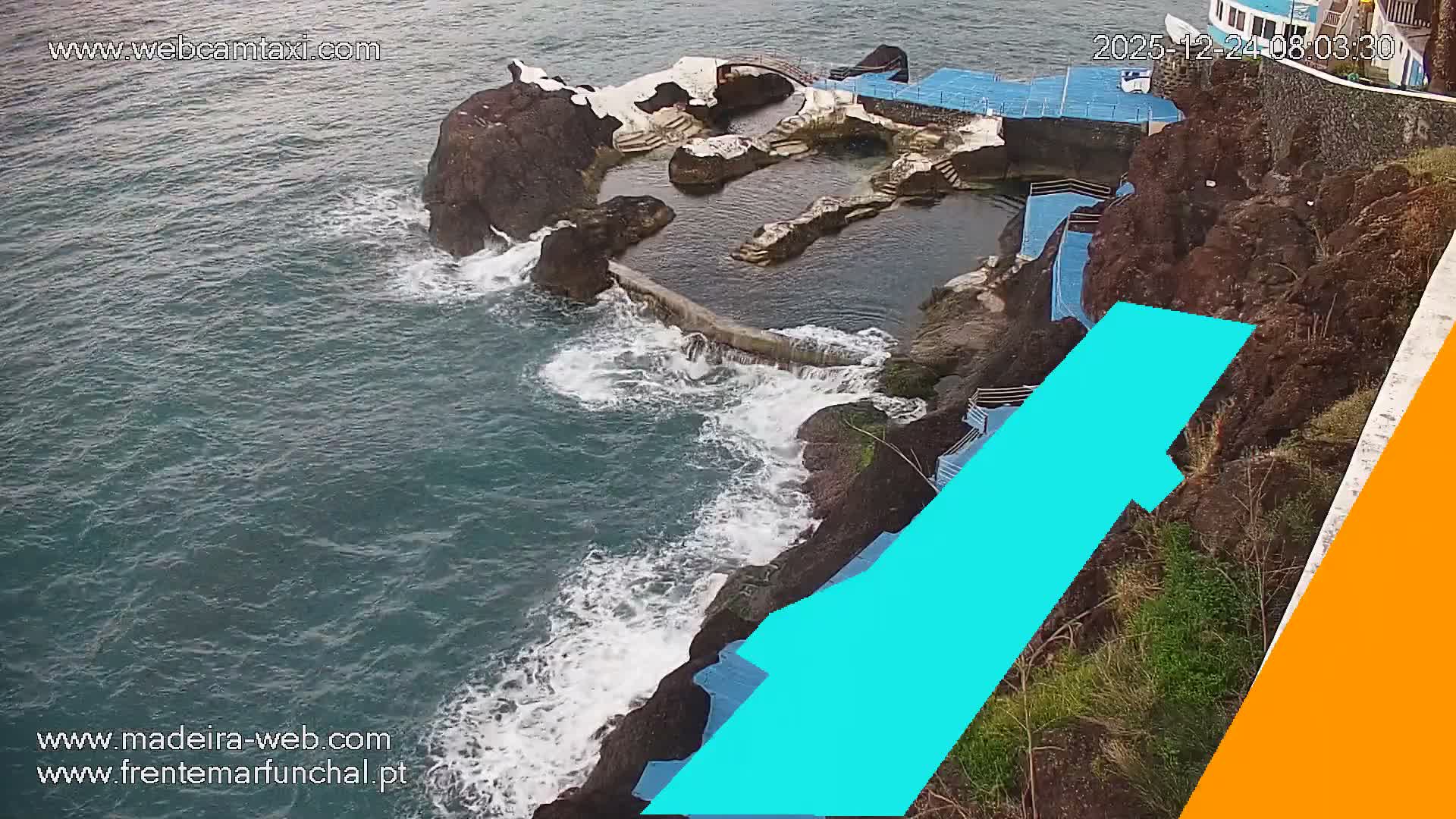Waves crash against a rocky coastline featuring natural sea pools and artificial structures built into the cliff, all beneath an overcast sky.