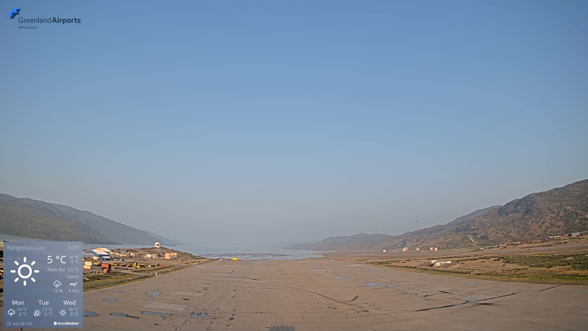 A sunny airport runway with a few puddles stretches out towards a calm body of water and low, brown hills under a clear, light blue sky.