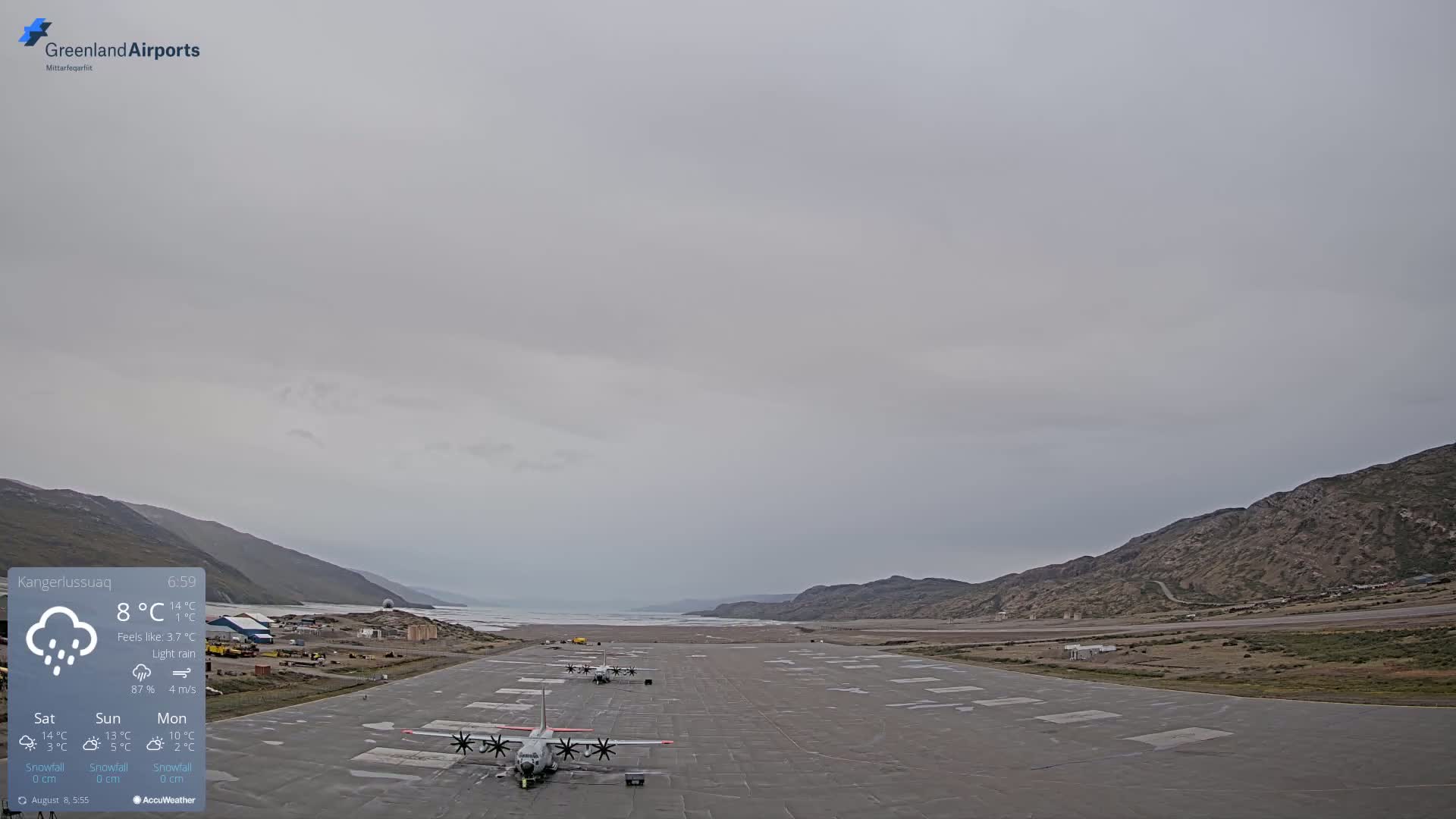A long airport runway with several propeller planes parked, under a light rain and overcast sky, is situated between a body of water and mountainous terrain.