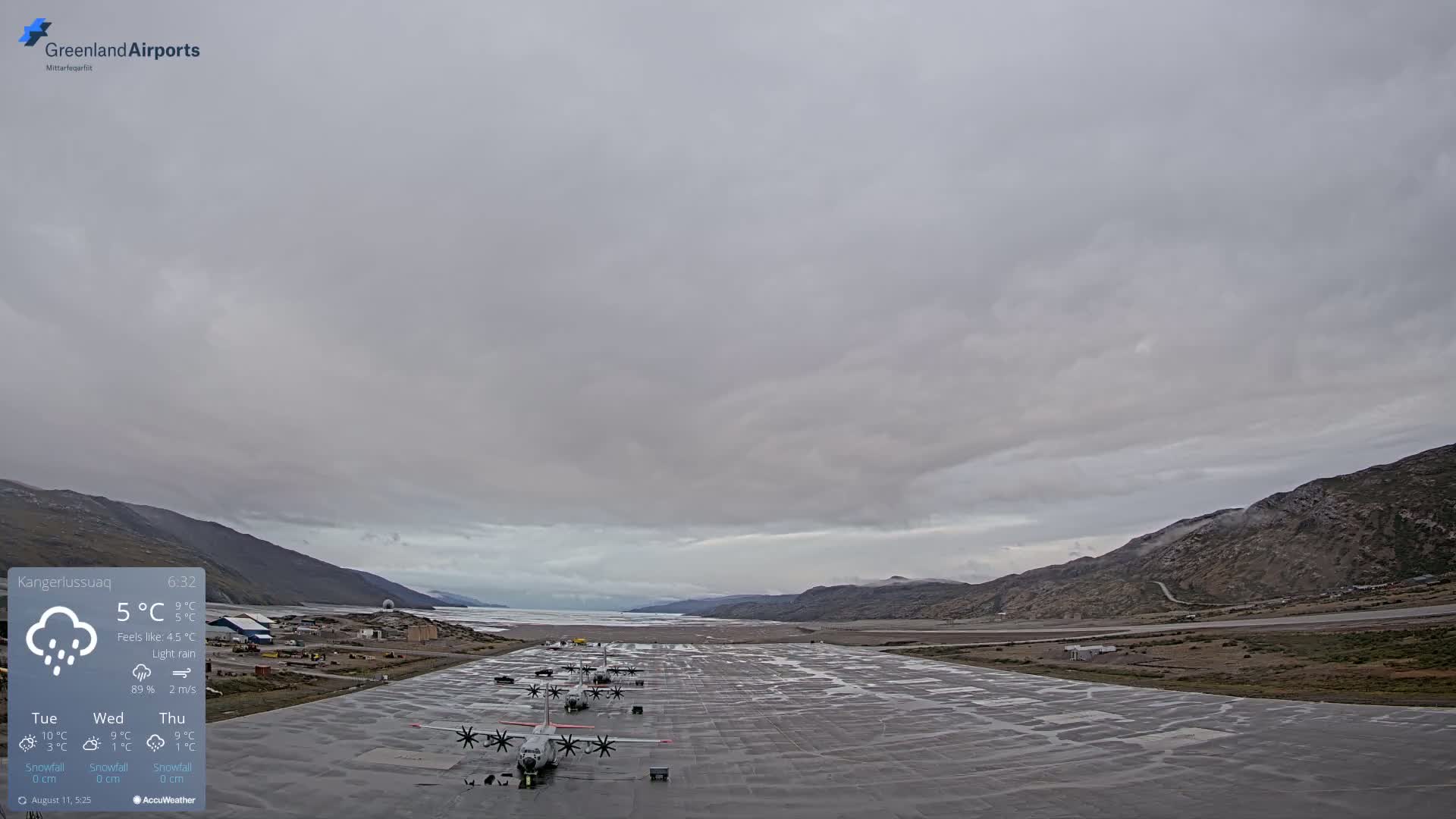 A wet airfield with several airplanes parked on it is surrounded by low mountains under a cloudy, overcast sky with light rain.