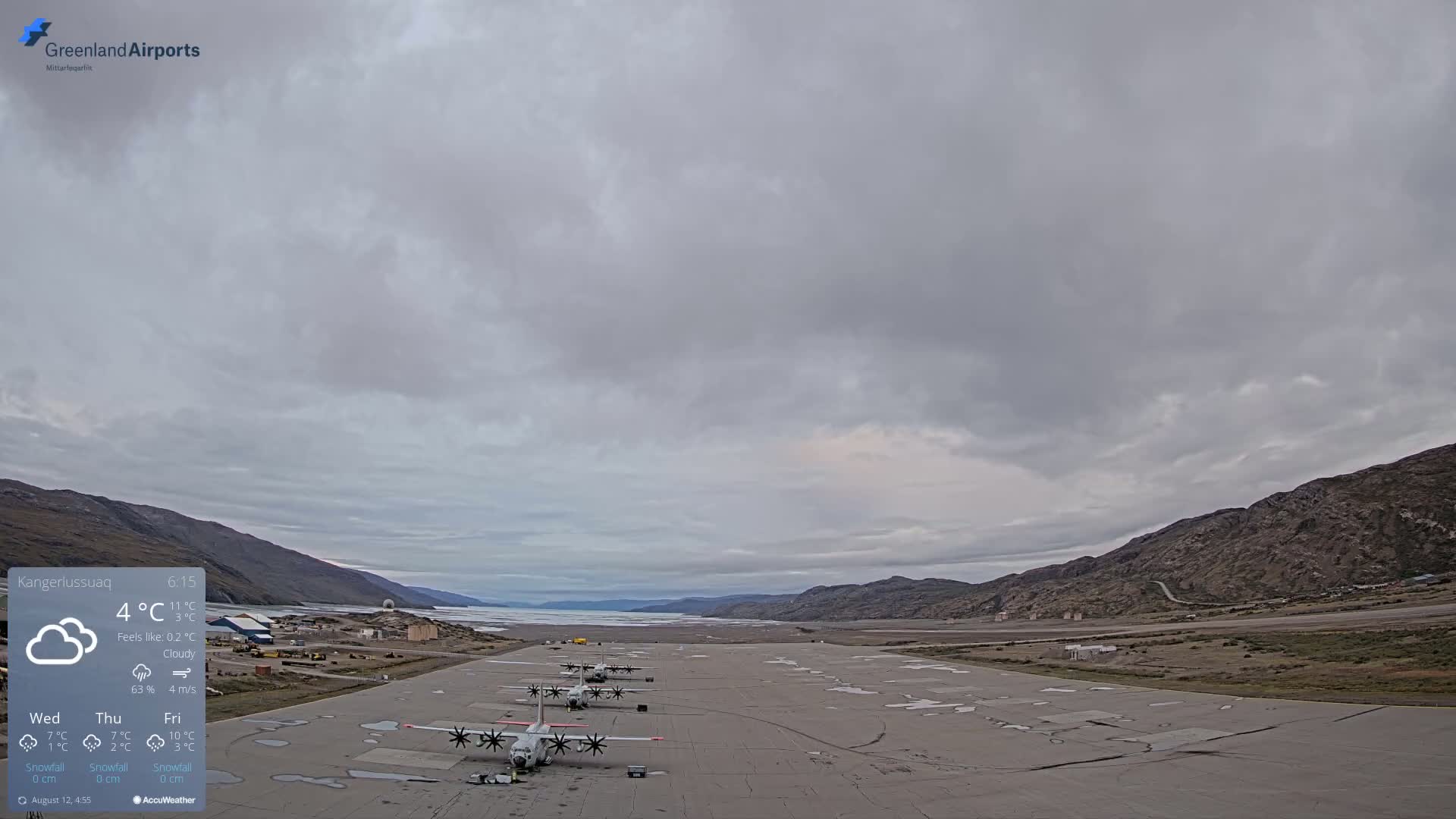 Several military transport planes are parked on a runway next to a body of water under a cloudy sky.